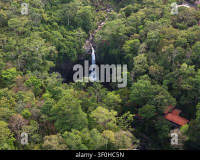Ein Wasserfall stürzt in ein Becken, das von dichtem Dschungel umgeben ist. Ein kleines Haus steht in der Nähe und ergänzt den üppigen Blick auf die Gegend. Stockfoto