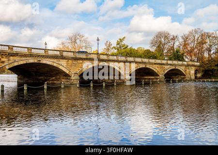Eine alte Steinbrücke mit weiten Bögen überspannt einen ruhigen See in London. Im Vordergrund verläuft eine Barriere aus Holzpfosten, die durch Ketten verbunden sind, entlang des Wass Stockfoto