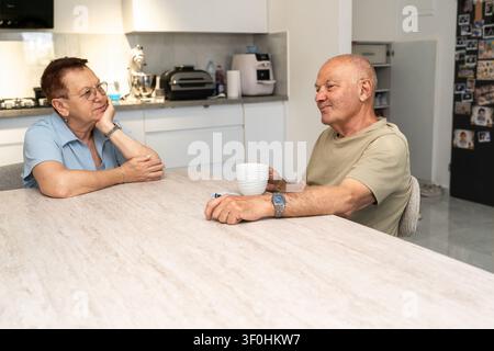 Zwei Senioren sitzen an einem hellen Tisch in einer Küche. Eine Person hält eine weiße Tasse. Sie genießen ein Gespräch in einer gemütlichen Umgebung. Stockfoto