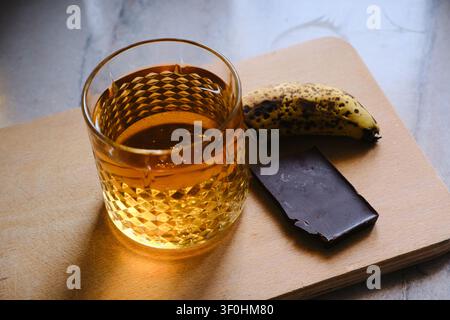 Glas goldener Whiskey mit Eiswürfeln, das auf einem dunklen Holztisch ruht. Die satte bernsteinfarbene Flüssigkeit reflektiert warmes Umgebungslicht und erzeugt sanfte Highlights Stockfoto