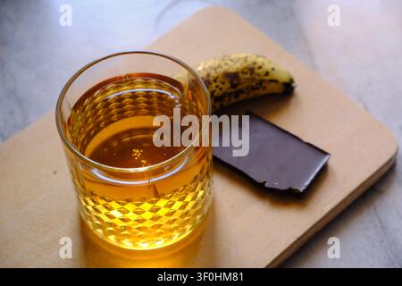 Glas goldener Whiskey mit Eiswürfeln, das auf einem dunklen Holztisch ruht. Die satte bernsteinfarbene Flüssigkeit reflektiert warmes Umgebungslicht und erzeugt sanfte Highlights Stockfoto