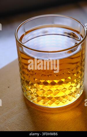 Glas goldener Whiskey mit Eiswürfeln, das auf einem dunklen Holztisch ruht. Die satte bernsteinfarbene Flüssigkeit reflektiert warmes Umgebungslicht und erzeugt sanfte Highlights Stockfoto