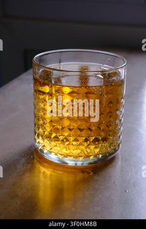 Glas goldener Whiskey mit Eiswürfeln, das auf einem dunklen Holztisch ruht. Die satte bernsteinfarbene Flüssigkeit reflektiert warmes Umgebungslicht und erzeugt sanfte Highlights Stockfoto