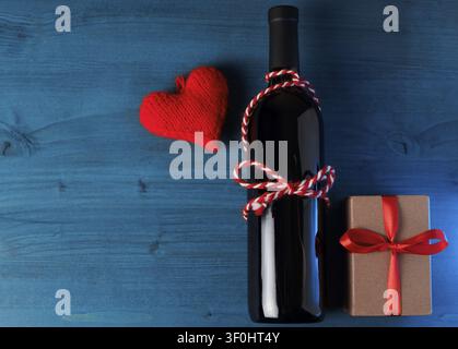 Eine Flasche Wein und ein Geschenk auf einem blauen Holztisch. Rotes gestricktes Herz, ein Symbol der Liebe und das Konzept des Valentinstags Stockfoto