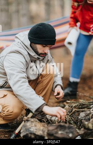 Freunde campen in einem Bergwald, während ein Mann ein Lagerfeuer neben einer Hängematte vorbereitet Stockfoto