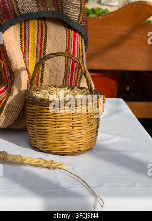 Vertikale Aufnahme eines kleinen geflochtenen Weidenkorbs gefüllt mit rohen Gerstengrannen neben einem gebundenen Weizenbündel und einem traditionellen gestreiften kretischen Textilsack. Stockfoto