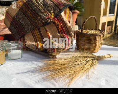 Alte Zea-Körner in einem Weidenkorb, Gewürzgläser und ein gebundener Strauß getrockneter Weizenähren neben einem traditionellen handgewebten kretischen Sack auf einer weißen Oberfläche Stockfoto