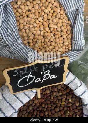 Kichererbsen und Linsen in gestreiften Säcken auf einer Holzoberfläche, versehen mit einem kleinen Schwarztafelschild mit dem griechischen Namen „Φάβα“ für die Linsen. Stockfoto