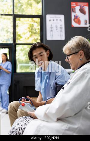 Kaukasische Ärztin überprüft den Blutdruck eines asiatischen männlichen Patienten während eines Standard-Klinikbesuchs. Junger erwachsener Mann lässt sich im Wartebereich des Krankenhauses einer routinemäßigen Gesundheitsuntersuchung unterziehen. Stockfoto