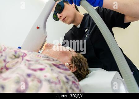 Seniorin liegt in einem Schönheitssalon-Bett, während eine Therapeutin eine Laser-Gesichtspflegeprozedur durchführt, Schutzbrille tragend, zur Verjüngung a Stockfoto