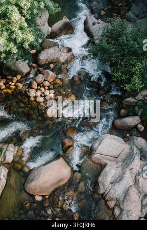 Luftaufnahme von klarem Flusswasser, das zwischen glatten Granitblöcken und runden Steinen rauscht, sich durch dichten grünen Wald windet mit weißen Stromschnellen und tr Stockfoto