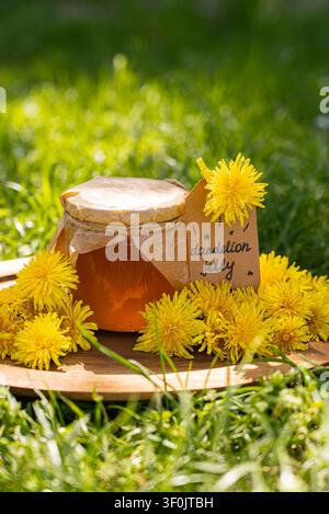 Hausgemachte Löwenzahn-Gelee in einem Gläser mit Papierdeckel und Anhänger, umgeben von frischen gelben Blüten auf einem Holzbrett im sonnigen Gras, hervorgehoben kannni Stockfoto