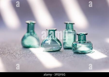 Vier kleine türkisfarbene Glasflaschen auf einer strukturierten Oberfläche, beleuchtet von sanftem Fensterlicht und Schatten. Minimales Stillleben mit geringer Schärfentiefe und Luft Stockfoto