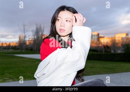Gen Z asiatische Frau, die im Abendlicht in einem Stadtpark posiert, ein rotes Hemd und einen weißen Mantel trägt, mit langen Haaren weg schaut, urbane Gebäude und stimmungsvolle Atmosphäre Stockfoto