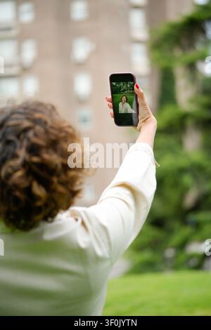 Eine Frau, die ein Smartphone hält und ein Selfie in einer grünen Parkumgebung macht. Ihr Fokus liegt auf dem Telefon, das ihr Spiegelbild auf dem Bildschirm einfängt, umgeben von Stockfoto