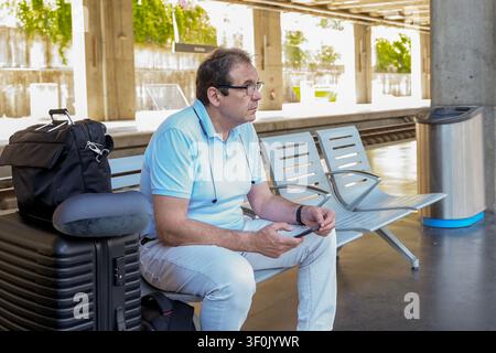 Ein reifer Mann in einem hellblauen Hemd wartet an einem Bahnhof, sitzt auf einer Metallbank mit seinem Koffer neben sich. Er überprüft sein Smartphone, bereit für Stockfoto