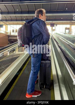 Ein reifer Reisender mit einem Rucksack und einem Koffer steht auf einer Rolltreppe in einem Bahnhof und blickt nach vorne. Sonnenlicht fällt durch große Fenster, hoch Stockfoto