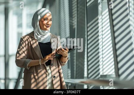 Selbstbewusste Geschäftsfrau mit Hijab steht in einem hellen modernen Büro mit gestreiften Fenster-Schatten, hält ein Tablet und plant Arbeit, Technologie und Stockfoto