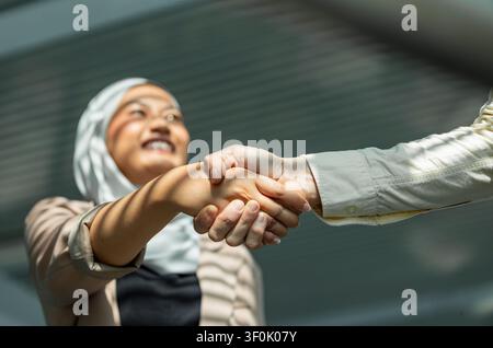 Nahaufnahme eines professionellen Händedrucks mit einer lächelnden muslimischen Frau im Hijab im Freien, der Vertrauen, Partnerschaft, Vielfalt und erfolgreiches Geschäft symbolisiert Stockfoto