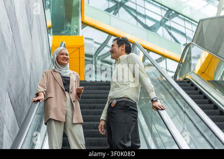Zwei asiatische Geschäftskollegen unterhalten sich, während sie eine Rolltreppe in einem hellen modernen Glasatrium mit schlanker Architektur, Reflexionen und einem urbanen Büro fahren Stockfoto