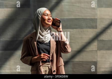 Selbstbewusste muslimische Geschäftsfrau mit Hijab telefoniert auf dem Smartphone in einer modernen Stadtszene, steht an einer Betonwand mit dramatischen Schatten, trägt Smart c Stockfoto