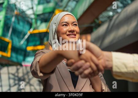 Nahaufnahme eines professionellen Händedrucks mit einer lächelnden asiatischen muslimischen Frau, die ein Hijab in einem modernen Büro-Atrium trägt, symbolisch für Partnerschaft, Vertrauen und Stockfoto