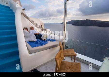 Seniorenpaar liegt auf einer Luxushotel-Terrasse in Santorini, Griechenland, und ruht auf einem Hängemattenbett mit Blick auf das Meer und die Caldera, blauen Treppen und ruhiger Sonne Stockfoto