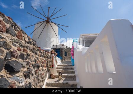 Reisende Frau geht mit ihrem Hund auf sonnigen Stufen in der Nähe einer traditionellen Windmühle und weiß getünchter Häuser auf einer griechischen Insel und fängt einen entspannten mediterranen Moment ein Stockfoto