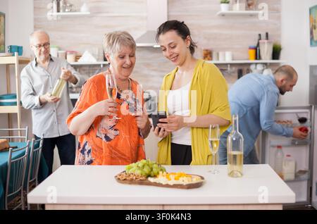 Mutter und Tochter nutzen ein Smartphone, um im Internet zu surfen, in einer Küche während eines Familientreffens mit Wein und Essen auf dem Tisch. Stockfoto