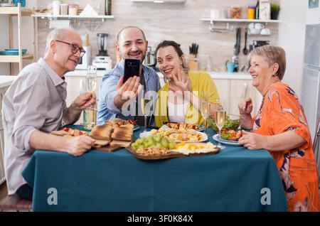 Fröhliche Familie genießt Brunch an einem Küchentisch während eines Videoanrufs, wobei die Eltern mit ihrem Sohn und dessen Frau in Kontakt treten, mit familiärer Bindung, Heimessen und gemeinsamer Kommunikation. Stockfoto