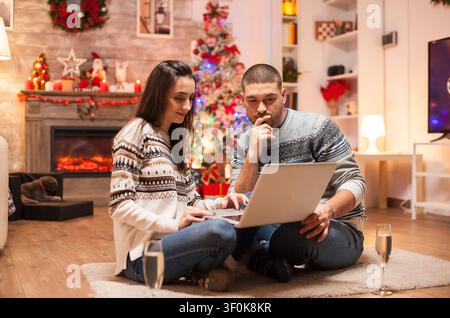 Paar in passender Kleidung genießt die Weihnachtsstimmung beim Surfen auf einem Laptop, entspannt zusammen in der Nähe festlicher Wohndekorationen mit warmen Details der Beziehung im Innenraum. Stockfoto