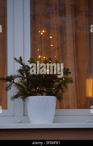 ein bezaubernder Miniatur-Weihnachtsbaum, geschmückt mit Lichterketten und roten Ornamenten, der auf einer Fensterbank in einem gemütlichen Wohninterieur ausgestellt ist, aufgenommen in einem Clo Stockfoto