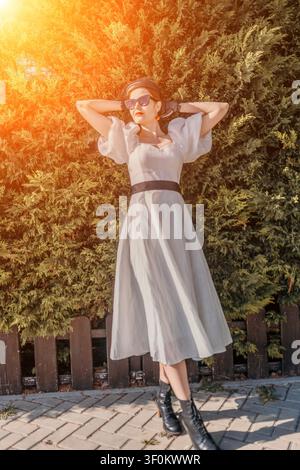 Frau im Kleid bei Sonnenuntergang posiert vor einer grünen Hecke und trägt eine Sonnenbrille und ein schwarzes Kopfstück Stockfoto