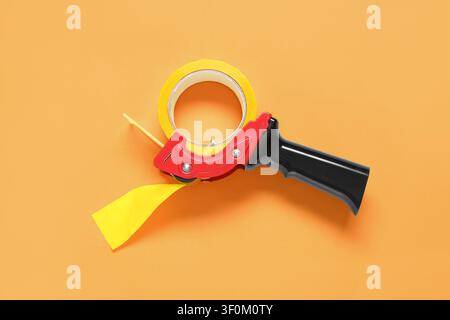Gelber Verpackungsbandspender mit einer Rolle Klebeband auf orangefarbenem Hintergrund, Ansicht von oben Stockfoto
