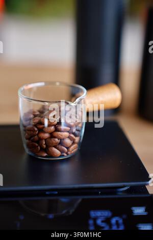 Geröstete Bohnen liegen in einer Glasbecher auf einer schwarzen Waage, wobei Kaffeewerkzeuge im Hintergrund unscharf sind. Konzept des Abwiegens von Kaffeebohnen, des Kaffeebrauens und der Rezeptzubereitung Stockfoto