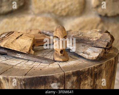 Kleine abstrakte Figur, handgeschnitzt aus Lorbeerholz auf einem Baumstamm, Nahaufnahme der traditionellen Schnitzerei in einer Handwerkswerkstatt neben Holzstücken und Werkzeugen. Stockfoto