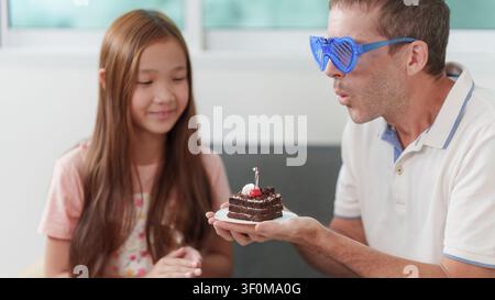 Lustiger Vater mit Herzbrille, der Kerze auf Geburtstagstorte mit Tochter ausbläst Stockfoto