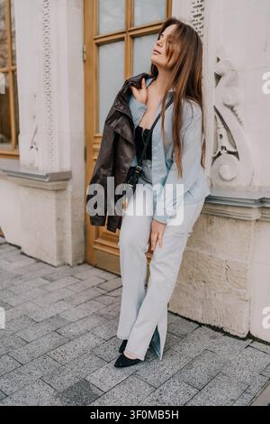 Modenporträt einer Frau, stilvolle Frau, die sich gegen ein weißes Steingebäude lehnt, ein blaues Hemd trägt und eine Lederjacke hält Stockfoto