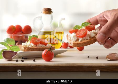 Hand, die einen knusprigen Toast hält, der mit Ricotta, Kirschtomaten und Basilikum belegt ist, umgeben von Olivenöl, Knoblauch und frischen Zutaten auf einem hellen Holztablett Stockfoto
