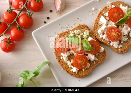 Zwei Vollkorntoasts mit zerbröckelter Feta, Kirschtomaten, Basilikum, Kräutern und Olivenöl auf einem weißen Teller, umgeben von frischen Tomaten, Knoblauch, Stockfoto