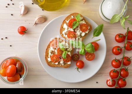 Oberansicht von zwei Toasts mit Ricotta und Kirschtomaten, garniert mit Basilikum auf einem weißen Teller, umgeben von Olivenöl, Knoblauch und frischem Gemüse auf Holz. Stockfoto