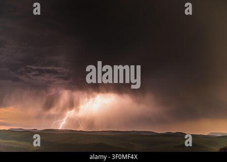 Ein mächtiger Blitzschlag erhellt einen sintflutartigen Regenschleier, der im Zwielicht über silhouettierte hügelige Ebenen hinabstürzt. Stockfoto