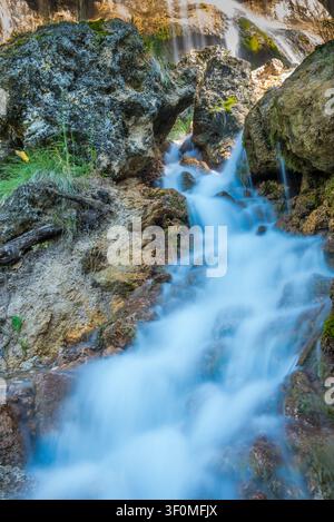 Ein mehrstufiger Bergwasserfall stürzt über moosbedeckte Kalksteinblöcke und felsige Ausläufer, aufgenommen mit einer Langzeitbelichtung, die r Stockfoto