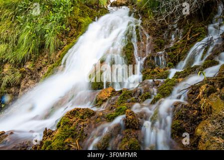 Ein mehrstufiger Kaskadenwasserfall stürzt über moosbedeckte vulkanische und sedimentäre Felsen, umgeben von dichter grüner Vegetation und freiliegenden Baumwurzeln Stockfoto