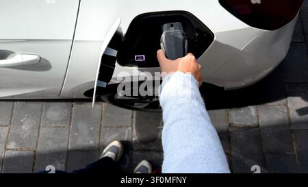 Eine Frau hält einen Ladestecker für Elektroautos in der Nähe des Ladensanschlusses eines weißen parkenden Autos Stockfoto