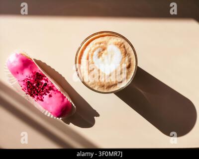 Pink eclair und ein Glas Cappuccino auf beigem Hintergrund in hellem Licht mit harten Schatten Stockfoto