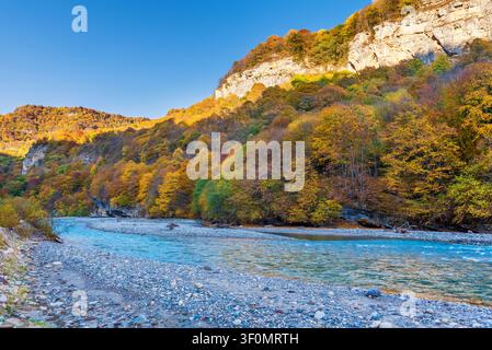 Ein türkiser Bergfluss windet sich durch ein felsiges Kiesufer unter hoch aufragenden Kalksteinhängen, die in leuchtendem Herbstlaub aus Gold und Orange gehüllt sind. Stockfoto
