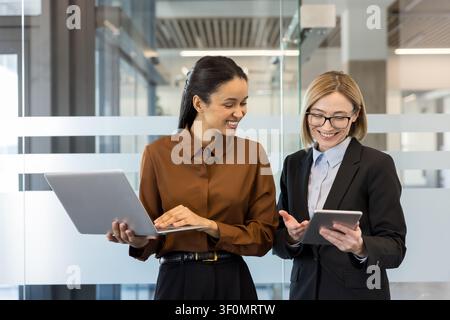 Zwei lächelnde multikulturelle Geschäftsfrauen, die zusammenarbeiten und Ideen diskutieren, während sie gemeinsam mit einem Laptop und einem digitalen Tablet in einer modernen Büroumgebung arbeiten Stockfoto