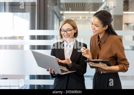 Zwei vielfältige Geschäftsfrauen, die aktiv zusammenarbeiten und lächeln, eine zeigt Inhalte auf einem Laptop, während die andere ein Klemmbrett hält, sie arbeiten zusammen in einer modernen Büroumgebung Stockfoto
