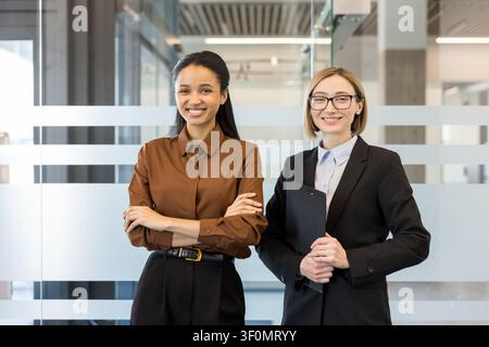 Zwei professionelle Geschäftsfrauen, eine mit verschränkten Armen und die andere mit einem Klemmbrett, stehen zusammen und lächeln in einem zeitgenössischen Büro und repräsentieren Teamarbeit und Erfolg Stockfoto